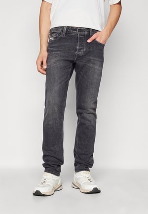Man draagt versleten zwarte slim-fit jeans en witte sneakers, staat met één hand in zijn zak tegen een witte achtergrond.