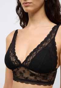 Musta pitsinen bralette, jossa on aaltoileva reuna. Siinä on läpikuultavia kukka-aiheisia yksityiskohtia, V-kaula-aukko ja säädettävät olkaimet. Karkea ja pehmeä materiaali.