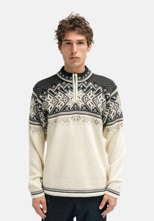 Ung mann iført en kremfarget og svart mønstret strikkegenser med halvglidelåsåpning ved kragen, stående foran en ensfarget lys grå bakgrunn.