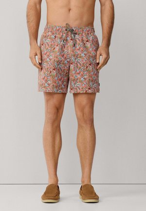 Man die koraalroze korte broek met bloemenprint en bruine instapschoenen draagt, staand tegen een effen lichtgrijze achtergrond.