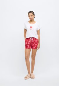 Λευκό κοντομάνικο μπλουζάκι με το κείμενο «AMOUR». Κόκκινα σόρτς με λευκό μοτίβο καρδιών, με ελαστική μέση και κορδόνι. Μπεζ παντόφλες.