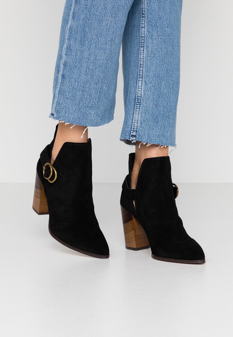 Botas de ante negras hasta el tobillo con punta afilada, tacón de madera grueso y detalle de hebilla lateral, combinadas con jeans de mezclilla azul con dobladillo deshilachado.