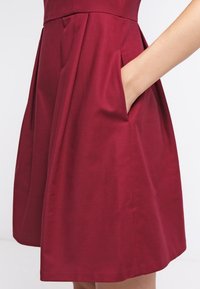 Main placée dans la poche latérale d'une robe rouge foncé plissée, sans manches et longueur genoux, sur un fond uni.