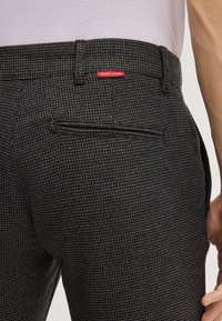 Nahaufnahme einer Person, die eine dunkle, gemusterte Hose trägt, mit einem kleinen roten "JOOP! JEANS"-Etikett über einer Gesäßtasche und einem in die Hose gesteckten weißen Hemd.