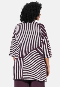 Donna con capelli ricci che indossa una maglietta geometrica a strisce viola e bianche, larga, e pantaloni abbinati, vista da dietro.