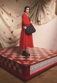 Borsa nera con motivo floreale colorato, maglione rosso e gonna in tulle su una piattaforma a scacchiera rosa e marrone con bordi decorativi.