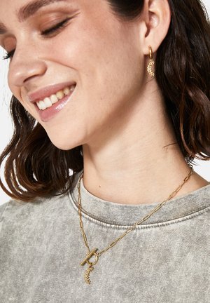 Guldtonet halskæde med en toggle-lås og en struktureret accent. Modellen bærer øreringe, der matcher halskædens design. Grå, afslappet beklædning.