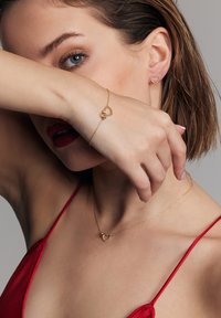 Złota biżuteria w kształcie serca, w tym kolczyki i bransoletka, prezentowana na modelce. Tło jest neutralne, z naciskiem na wzornictwo biżuterii.