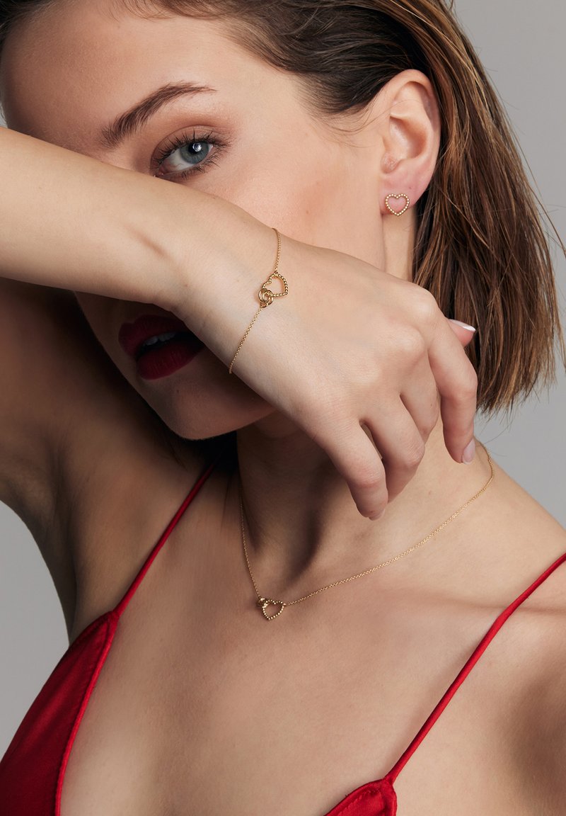 Złota biżuteria w kształcie serca, w tym kolczyki i bransoletka, prezentowana na modelce. Tło jest neutralne, z naciskiem na wzornictwo biżuterii.