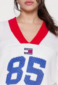 Bílý pletený svetr s červeným vroubkovaným véčkovým výstřihem, na kterém se nachází modré číslo "85" a logo "TOMMY JEANS" na hrudi. Texturovaný design.