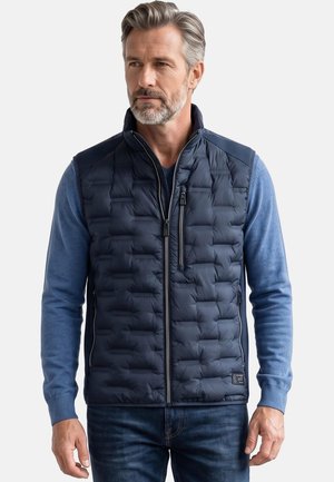 Mann mit grauen Haaren und Bart, der eine marineblaue gesteppte Weste über einem blauen Langarmhemd und blauen Jeans trägt, steht vor einem einfarbigen Hintergrund.