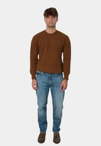 Maglione marrone a coste con maniche lunghe, abbinato a jeans blu e scarpe marroni. Il modello è in piedi contro uno sfondo bianco semplice.