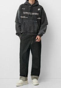 Sudadera con capucha negra de gran tamaño con impresiones de texto, combinada con pantalones anchos de mezclilla oscura. Las zapatillas son blancas con detalles en gris claro.