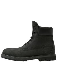 Des bottines en suède noir présentent un col rembourré, un laçage à six œillets et une semelle en caoutchouc texturée, avec un logo sur le côté.