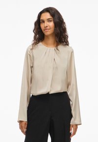 Μπεζ μπλούζα με μακριά μανίκια και στρογγυλή λαιμόκοψη, με πτυχώσεις μπροστά, φτιαγμένη από μαλακό, μεταξένιο ύφασμα. Φορεμένη με μαύρο παντελόνι.