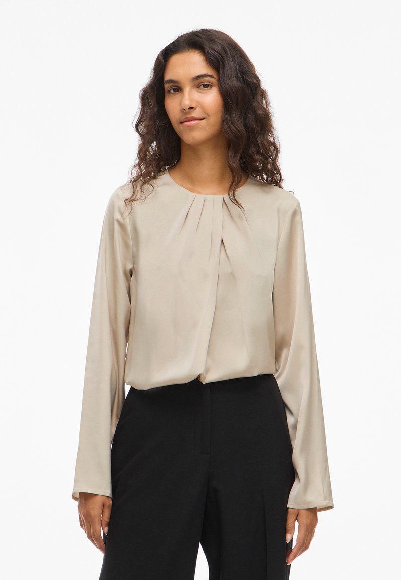 Μπεζ μπλούζα με μακριά μανίκια και στρογγυλή λαιμόκοψη, με πτυχώσεις μπροστά, φτιαγμένη από μαλακό, μεταξένιο ύφασμα. Φορεμένη με μαύρο παντελόνι.