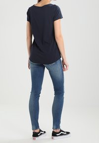 Žena stojící vzadu, oblečená v černém tričku, modrých úzkých džínách a černo-bílých teniskách na světlém jednolitém pozadí.