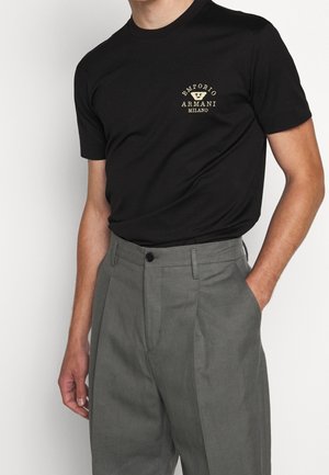 Mężczyzna w czarnej koszulce Emporio Armani Milano, włożonej w wysokie szare spodnie z plisami, zapinane na guzik i z szlufkami na pasek.