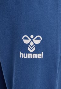 Blauer Stoff mit einer strukturierten, gestrickten Oberfläche. Verfügt über ein weißes Logo mit abstrakten Formen und dem Wort "hummel" darunter.