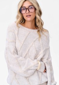 Kobieta z blond włosami, nosząca za dużą oprawkę w stylu żółwi oraz fakturowany, off-white sweter przeciwko białemu tłu.