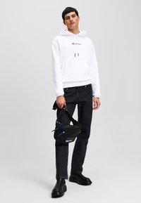Weißer Hoodie mit Logo, schwarze Jeans und schwarze klobige Stiefel. Schwarze Umhängetasche mit blauen Akzenten. Einfaches Design, einfarbige Farben.