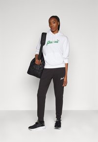 Sudadera blanca con el logo verde de "Puma", pantalones de chándal negros con logo, zapatos negros con detalles verdes y una mochila negra. Tejido suave, corte relajado.