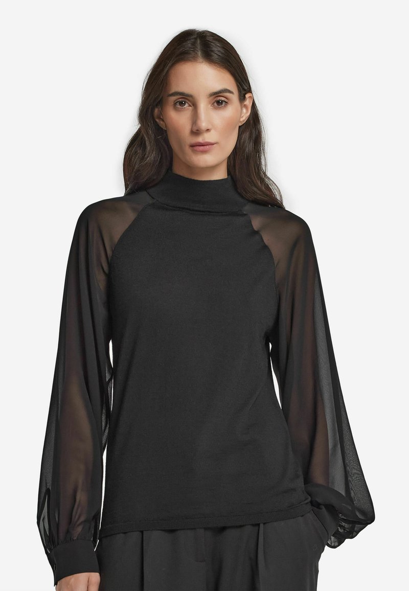 Top nero con corpo solido e maniche lunghe raglan trasparenti, colletto alto e maniche con polsini. Texture morbida, design minimale.