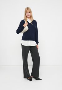 Navy v-neck sweater layered over a white shirt, paired with gray pinstripe wide-leg trousers and black pointed-toe mules. Holding a cream handbag.