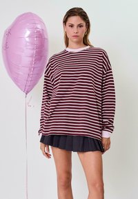 Bordovai ir baltai dryžuota ilgomis rankovėmis marškinėliai su apvalia iškirpte, poruojami su tamsiai raukšlėta sijonu. Šalia yra rožinis širdelės formos balionas.