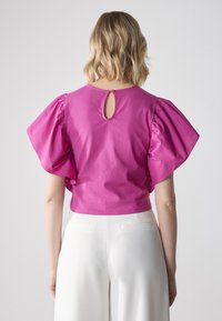 Fuchsia Top mit Puffärmeln und einem Schlüsselmodus im Rücken, aus leichtem Material. Kombiniert mit weißen Hosen, die zarte Taschenakzente aufweisen.