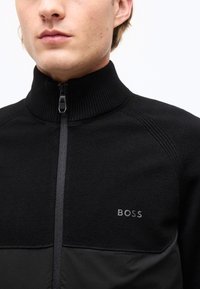Homme portant un pull noir à fermeture éclair avec un col haut côtelé et le logo "BOSS" sur la poitrine, sur fond blanc.