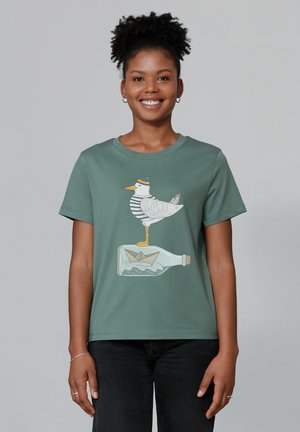 Olijfgroen t-shirt met een afbeelding van een meeuw in een gestreept shirt die op een fles staat met een papieren bootje erin. Eenvoudig ontwerp met ronde hals.