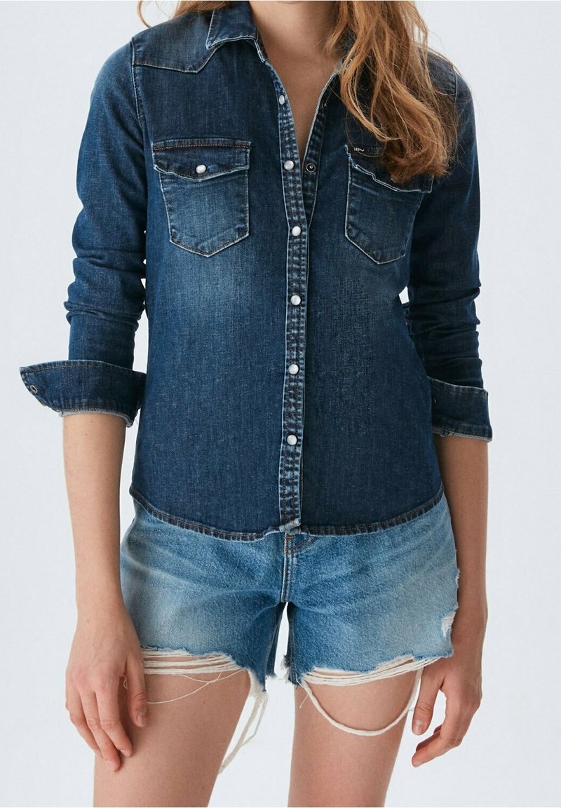 Personne portant une chemise en jean foncé à boutons avec des manches retroussées et un short en jean bleu clair usé aux bords effilochés.