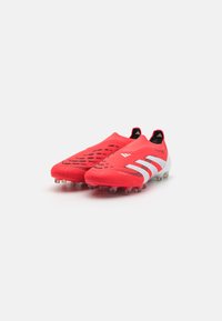 Bottines de football rouges avec une tige texturée, présentant un motif en nid d'abeille, des rayures blanches sur le côté et des crampons transparents sur la semelle.