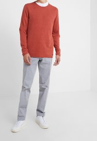 Långärmad t-shirt i rostfärg med rund halsringning, kombinerad med ljusgrå byxor och vita sneakers. Har en minimalistisk design.