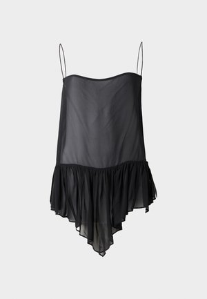 Femme aux longs cheveux foncés porte un top noir transparent sans manches avec un ourlet à volants, un pantalon blanc à jambes larges et des chaussures noires ouvertes à l'avant, debout de manière neutre.