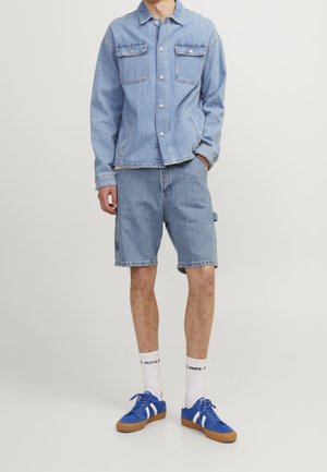 Mand iført en lys blå denimjakke og matchende denimshorts, hvide sokker og blå sneakers med hvide striber.