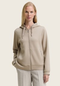 Beige zip-up hoodie met een capuchon, trekkoorden en voorzakken. Zachte stof, aansluitende boorden en zoom. Gedragen over lichtgrijze broeken.
