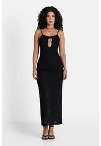 Schwarzes Maxikleid mit taillierter Passform, das über einen zentralen Schlüssel-Ausschnitt und dünne Träger verfügt. Texturiertes Material, knöchellang, kombiniert mit Riemchensandalen.