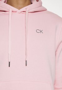 Svetloružová mikina s kapucňou a šnúrkou, predným vreckom a malým vyšitým logom "CK" na hrudi. Hladká textúra, neformálny dizajn.