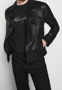 Black leather jacket with snap closures, two front pockets, and a classic design. Worn over a dark sweater, paired with dark jeans.
