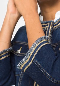 Jeansová bunda s roztrženými manžetami, modré barvy, se zlatými švy a detaily na knoflících. Detailní pohled na texturu a design rukávu.