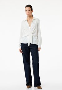 Witte blouse met een knoopdetail aan de voorkant, gecombineerd met donkerblauwe flared jeans en beige puntige schoenen. Eenvoudig ontwerp, lichte textuur.