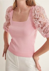 Haut en maille rose avec un décolleté carré et des manches bouffantes en dentelle ornées de motifs floraux. Bordures côtelées à l'ourlet et aux poignets ajoutant de la texture.