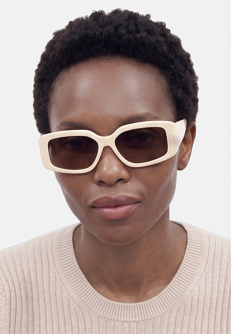 Femme aux cheveux courts et bouclés portant des lunettes de soleil rectangulaires beige clair et un pull côtelé beige sur un fond uni.