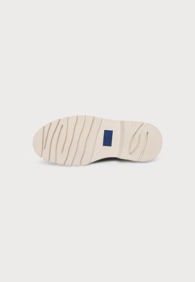 Semelle de chaussure beige clair avec des motifs de bande de roulement ondulés et une petite étiquette rectangulaire bleu foncé au centre, présentée sur un fond blanc.