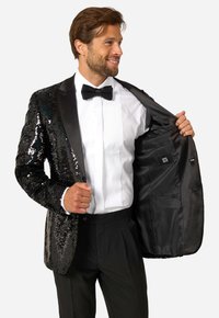 Czarna smokingowa marynarka z cekinami i satynowymi klapami, noszona na białej plisowanej koszuli i czarnej muszce, z widocznym detalem wewnętrznej kieszeni.