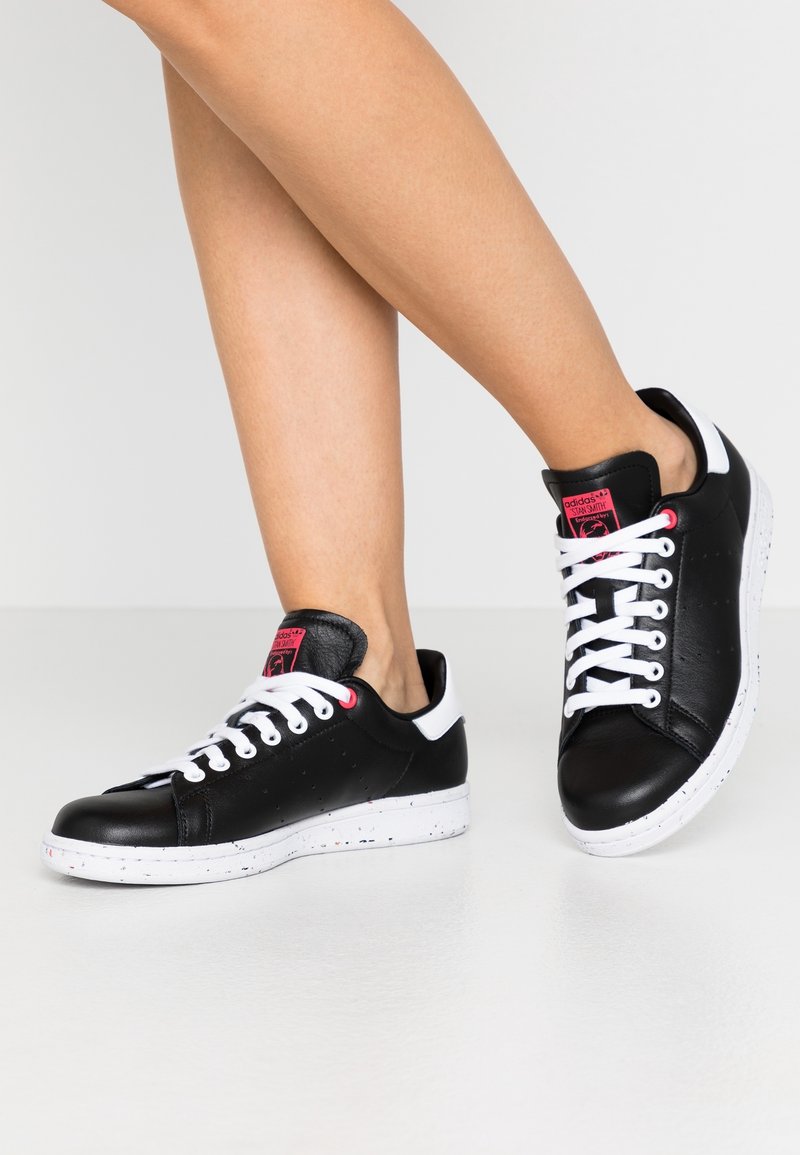 Svarta lädersneakers med vita accenter, rund tå, perforerade detaljer, vita snörningar och en texturerad gummisula med färgglada prickar.