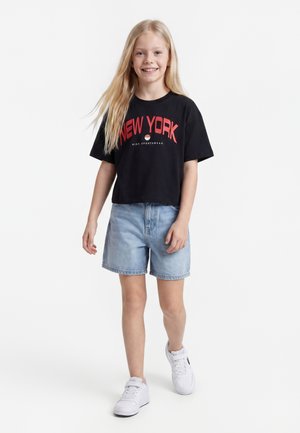 Młoda dziewczyna z długimi blond włosami, ubrana w czarną koszulkę New York, jasnoniebieskie jeansowe szorty i białe trampki, idzie i uśmiecha się do przodu.