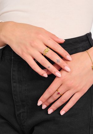 Hände mit hellrosa Nägeln, kleinen schwarzen Fingertattoos, die goldene Ringe mit einem roten Edelstein und einem floralen Design tragen, ruhend auf schwarzer Hose.